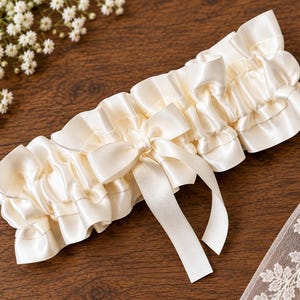 May include: An ivory satin garter featuring a gathered, ruffled design with a central decorative bow. The accessory is constructed from smooth, lustrous fabric with a consistent cream-colored tone throughout the entire piece, including the ribbon tails.