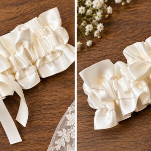 May include: An ivory satin garter featuring a gathered, ruffled design and a central satin ribbon bow with two hanging tails. The accessory is constructed from smooth, lustrous fabric and displays a consistent, pleated texture throughout its length.