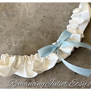 May include: White satin garter with a light blue satin bow. The garter has a ruffled edge and is tied with a ribbon.