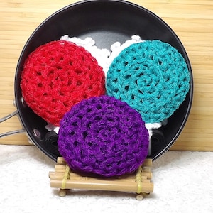 May include: Three crocheted hair scrunchies in a black pan. The scrunchies are red, teal, and purple.