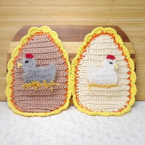 May include: Two crocheted egg-shaped pot holders with a chicken design. One pot holder is brown with a gray chicken and the other is white with a white chicken.