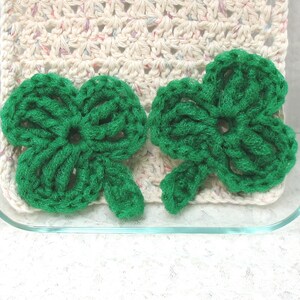 Op de afbeelding: Twee felgroene gehaakte klavers liggen op een crèmekleurige gehaakte pannenlap. De pannenlap zit in een doorzichtige glazen ovenschaal. De klavers hebben drie bladeren en een steel, perfect voor St. Patrick's Day.