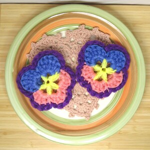 Puede incluir: Dos flores de ganchillo de color morado y rosa con centros amarillos en un plato marrón y verde. Las flores están hechas con una variedad de puntos y colores.