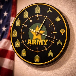 May include: A round black wall clock with gold-colored hands and numerals. The clock face features the word "ARMY" in gold, a gold star, and military rank insignias. The clock has a gold-colored border and is mounted on a beige wall.