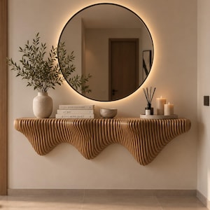 Puede incluir: Una mesa consola montada en la pared con un diseño de listones de madera ondulados. Encima cuelga un espejo redondo grande con retroiluminación. La mesa sostiene un jarrón blanco con vegetación, tres libros, un cuenco, un difusor y dos velas.