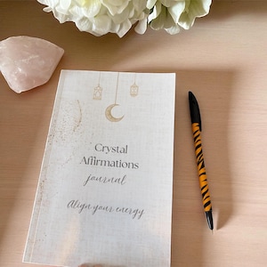 Op de afbeelding: Een dagboek met de tekst "Crystal Affirmations journal Align your energy" op de omslag, met een halve maan en lantaarn ontwerp. Daarnaast ligt een pen met een oranje en zwart tijgerstreep patroon. Een rozenkwartskristal en witte bloemen op de achtergrond.