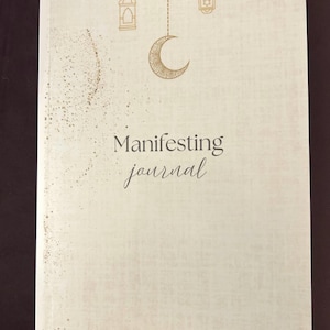 May include: A cream-colored journal with the words "Manifesting journal" in elegant script. The cover features gold illustrations of a crescent moon, lanterns, and scattered dots. The journal is closed and appears to be new.