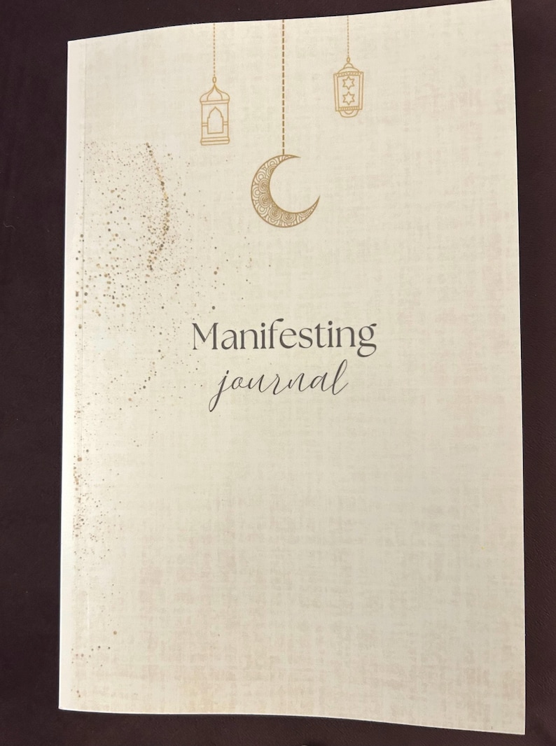 May include: A cream-colored journal with the words "Manifesting journal" in elegant script. The cover features gold illustrations of a crescent moon, lanterns, and scattered dots. The journal is closed and appears to be new.