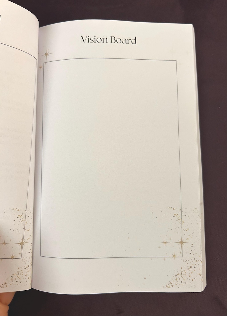 May include: Open notebook with the words "Vision Board" at the top. The blank page is framed with a thin black border. Gold starburst and speckle designs are in the corners of the page.