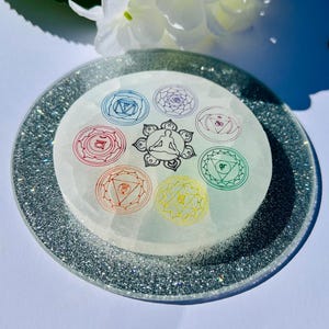 May include: A round, white crystal plate with a silver glitter rim. The center features a detailed illustration of a person in a seated yoga pose, surrounded by seven colorful chakra symbols. A spiritual and decorative item.