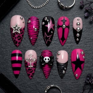 May include: A set of ten almond-shaped press-on nails featuring a pink and black color palette. Designs include leopard print, zebra stripes, horizontal stripes, silver star charms, a cross emblem, a skull with a bow, and silver studs.