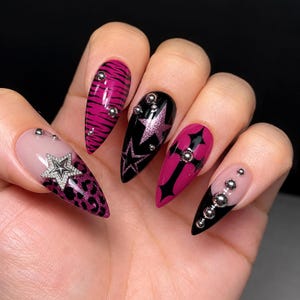 May include: Almond-shaped nails featuring a mix of designs including pink tiger stripes, black with pink glitter stars, a pink cross, and a nude base with leopard print and a silver star charm. Each nail is accented with small, round silver metal beads.
