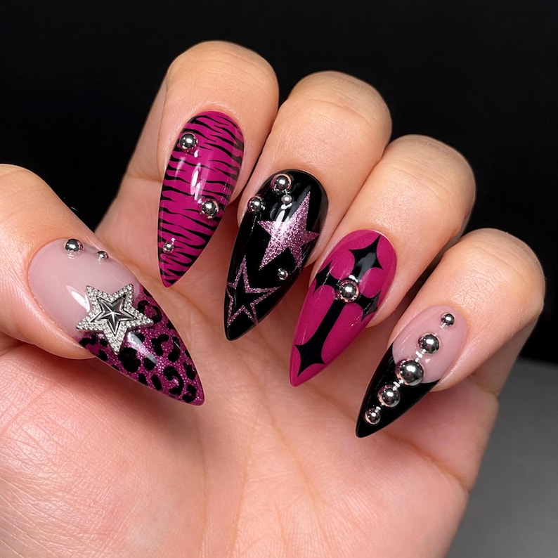 May include: Almond-shaped nails featuring a mix of designs including pink tiger stripes, black with pink glitter stars, a pink cross, and a nude base with leopard print and a silver star charm. Each nail is accented with small, round silver metal beads.