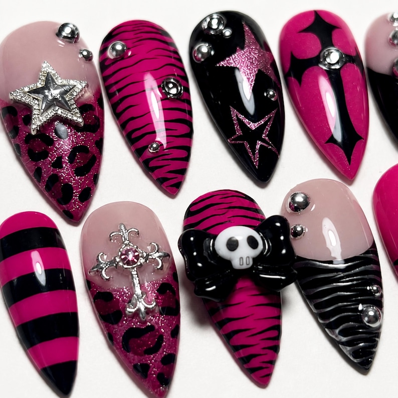 May include: A set of almond-shaped press-on nails featuring a variety of designs including pink and black leopard print, zebra stripes, horizontal stripes, and solid black. Embellishments include silver star charms, metal crosses, and a skull.