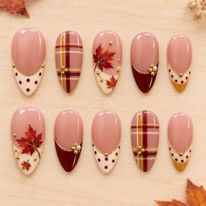 May include: A set of ten almond-shaped press-on nails featuring autumn-themed designs. The collection includes patterns with red maple leaves, burgundy and mustard yellow tips, plaid motifs, and white polka dots, accented with small gold beads.