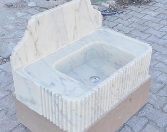 Può includere: Un lavabo in marmo bianco con un bordo decorativo scanalato e uno schienale. Il lavabo è montato su una base in legno.