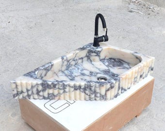 Può includere: Un lavandino in marmo bianco e viola con un rubinetto nero. Il lavandino è su una base di legno e ha un bordo ondulato.