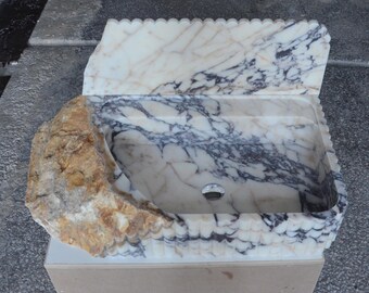 May include: A white marble sink with a unique design. The sink is carved to resemble a rock formation with a large, rounded stone on one side. The sink has a drain hole and is set on a beige base.