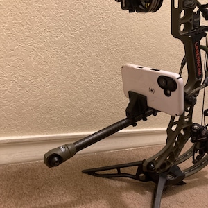 May include: A compound bow with a white smartphone attached. The bow is olive green with black accents and a sight. The smartphone is in a white case. The bow is resting on a black stand.
