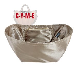 Könnte beinhalten: Ein beiger Satin-Handtaschen-Organizer mit einer kleinen Reißverschlusstasche im Inneren. Der Organizer hat eine rechteckige Form und ist für die Verwendung in einer Handtasche konzipiert. Der Text "Made For C-Y-M-E" ist sichtbar.