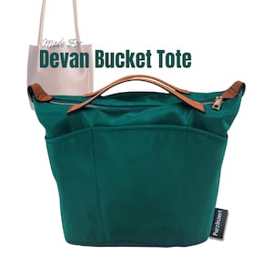 Puede incluir: Un bolso tote tipo cubo de color verde azulado con asas de cuero marrón y cierre de cremallera. El bolso tiene múltiples bolsillos exteriores y el texto "Devan Bucket Tote" es visible. En el fondo, un bolso tote más pequeño y de color claro.