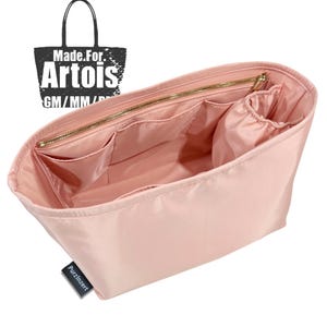 Pode incluir: Um organizador de bolsa rosa claro com vários bolsos e fecho de correr. O organizador tem formato retangular e é feito de um material liso, possivelmente nylon. O texto "Made For. Artois" é visível no canto superior esquerdo.