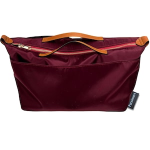 May include: A burgundy tote bag with a gold zipper and tan leather handles. The bag has a rectangular shape with a red interior and a small tag that reads "Purzinzert". The bag is made of a smooth, shiny material.