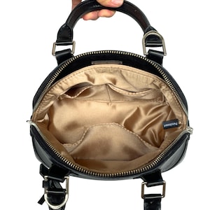 May include: Black handbag with a rounded shape, featuring a top handle and silver-tone hardware. The interior is lined with a gold satin fabric and includes multiple pockets. The bag is held by a hand.