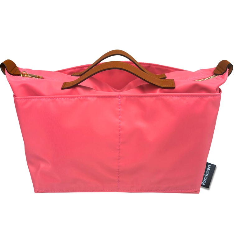 May include: A coral-colored tote bag with brown leather handles and gold zippers. The bag has two exterior pockets and a rectangular shape. The brand name "Purzinzert" is on a small black tag.