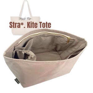May include: Beige tote bag interior organizer with multiple pockets and a zippered top. The bag has a light beige exterior and a gold-colored zipper. The text "Made For Stra*. Kite Tote" is visible in the image.