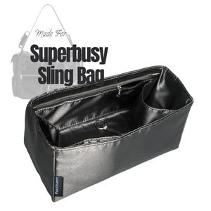 Peut inclure: Un organisateur de sac rectangulaire noir avec plusieurs compartiments intérieurs, une poche zippée et un anneau porte-clés central en métal. Le texte Made For SUPERBUSY Sling Bag est visible au-dessus, avec une étiquette PurzInzert sur le côté.