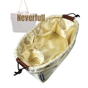 Op de afbeelding: Crèmekleurige tassenorganizer met meerdere compartimenten en een trekkoordsluiting. De binnenkant is van lichtgeel satijnachtig materiaal. De tas heeft bruine lederen accenten en de tekst "Made For Neverfull".