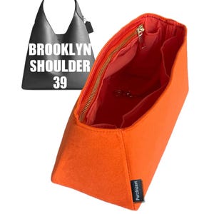 Könnte beinhalten: Ein orangefarbener Filz-Taschenorganizer mit goldenem Reißverschluss und mehreren Innentaschen. Die Tasche ist offen und zeigt einen leuchtend orangefarbenen Innenraum. Eine schwarze Umhängetasche mit dem Text "BROOKLYN SHOULDER 39" befindet sich im Hintergrund.