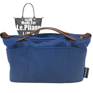 Puede incluir: Organizador de bolso azul con asas y tiradores de cremallera de cuero marrón. El organizador tiene múltiples bolsillos y un cierre de cremallera. El texto "Made For Le Pliage" es visible en el fondo.