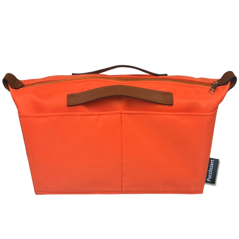 May include: Bright orange handbag organizer with brown leather handles and trim. The organizer has a zippered top and multiple pockets. The brand name "PurzInzert" is printed on a small tag.