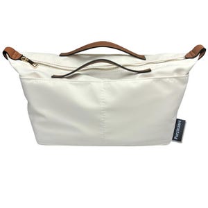 May include: Off-white handbag with brown leather handles and gold-colored zippers. The bag has a rectangular shape with a top zipper closure and a small tag that reads "Purzinzert". The bag is made of a smooth, possibly nylon, material.
