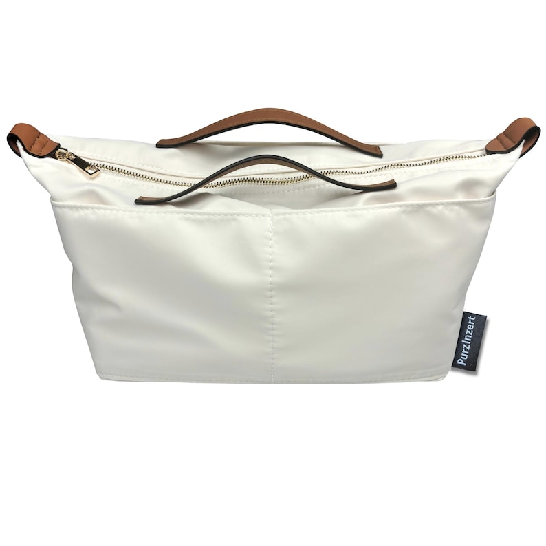 May include: Off-white handbag with brown leather handles and gold-colored zippers. The bag has a rectangular shape with a top zipper closure and a small tag that reads "Purzinzert". The bag is made of a smooth, possibly nylon, material.