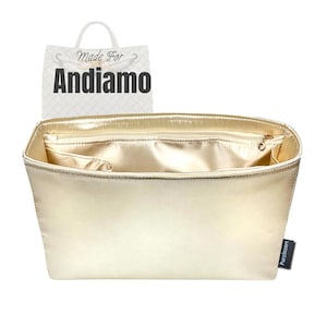 May include: A gold-colored handbag organizer with multiple compartments and a zippered top. The organizer has a satin-like finish and a small tag that says "Purzinzert". A shopping bag with the words "Made For Andiamo" is in the background.