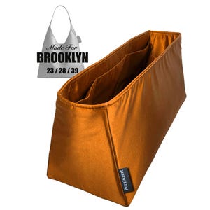 May include: A burnt orange handbag organiser with several internal pockets. The rectangular organiser is made from a smooth, shiny material. The text "Made For BROOKLYN 23/28/39" is visible in the image.