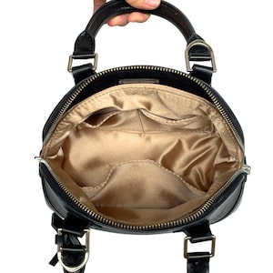 May include: Black, round handbag with a top handle and silver-tone hardware. The interior features a gold satin lining with multiple pockets. The bag has a zipper closure and a small bow detail on the front.