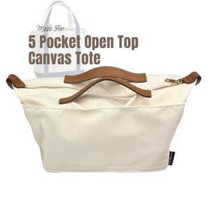 Puede incluir: Bolso tote de lona color crema con asas de cuero marrón y cremallera dorada. La bolsa tiene cinco bolsillos y el texto "5 Pocket Open Top Canvas Tote". Un contorno gris claro de un bolso tote está en el fondo.