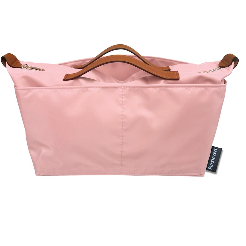 May include: A light pink tote bag with brown leather handles and a zipper closure. The bag has a rectangular shape and a small tag that reads "PurzInzert". The bag is made of a smooth, possibly nylon, material.