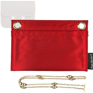 May include: A red satin bag with gold grommets and a gold chain necklace. The bag has a small black tag with the word "PurzInzert" printed on it. A white leather bag is in the background.