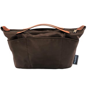 May include: A brown nylon handbag with a top handle and a zipper closure. The bag has two exterior pockets and a brown leather handle. The bag has the text "Purzinzert" on the side.