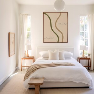 May include: A bedroom with a white bed, pillows, and a beige throw blanket. A wooden bench sits at the foot of the bed. A framed art piece with the text "one thing at a time" hangs above the bed. Two small wooden side tables with lamps are on either side of the bed.