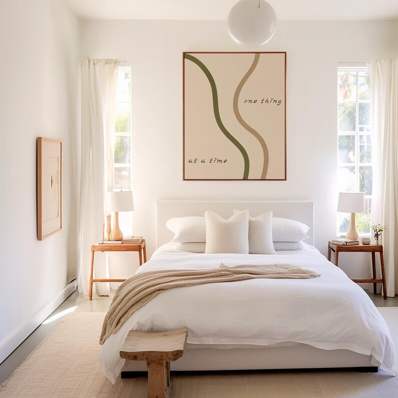 May include: A bedroom with a white bed, pillows, and a beige throw blanket. A wooden bench sits at the foot of the bed. A framed art piece with the text "one thing at a time" hangs above the bed. Two small wooden side tables with lamps are on either side of the bed.