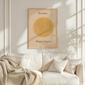 Può includere: Una stampa incorniciata con le parole "Breathe." e "Begin Again." in nero. L'opera presenta un grande cerchio giallo su sfondo beige. La cornice è di colore marrone chiaro. La stampa è appesa sopra un divano bianco con cuscini e una coperta.