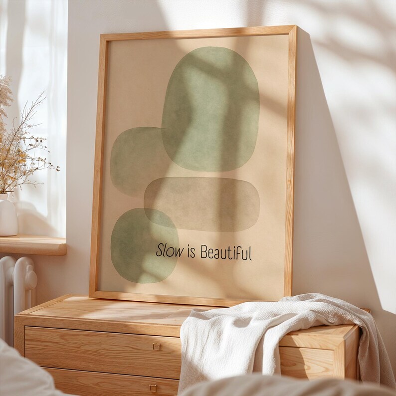May include: A framed art print featuring abstract green shapes on a beige background. The text "Slow is Beautiful" is printed in black. The artwork is displayed on a wooden dresser.