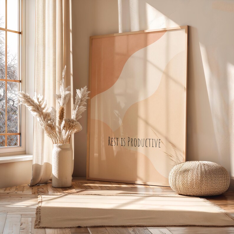May include: A framed art print with abstract shapes in peach and cream tones, with the text "REST IS PRODUCTIVE." The artwork leans against a wall in a room with a neutral color palette, a woven pouf, and a vase of dried flowers.