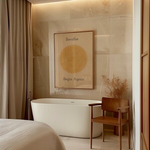May include: A modern bathroom scene featuring a white freestanding bathtub, a wooden chair, and a framed art print. The art print has the words "Breathe. Begin Again." and a yellow circle. Beige curtains and tile complete the spa-like aesthetic.